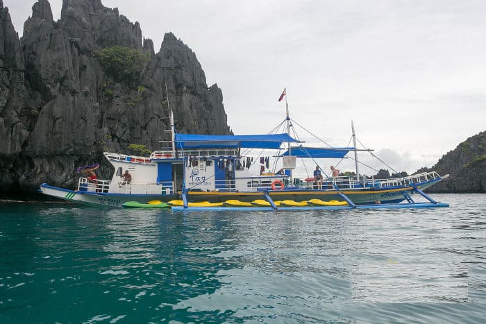 A blue and white outrigger boat with a blue canopy floats on calm turquoise waters near rugged rocky cliffs under a cloudy sky, capturing the diverse sizes of beauty in the Philippines. Several people are visible on the boat, with lush green vegetation atop the cliffs.