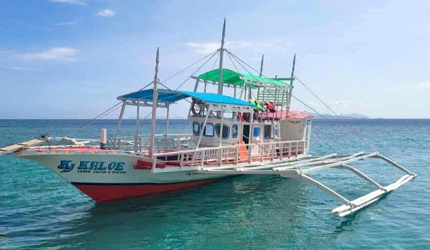 A traditional outrigger boat with colorful canopies is docked on a calm, clear ocean, perfect for island hopping. The vessel boasts a white and red hull with "Kiloe" labeled on the side. The sky is clear and blue, with distant land visible on the horizon, capturing the essence of the Philippines.