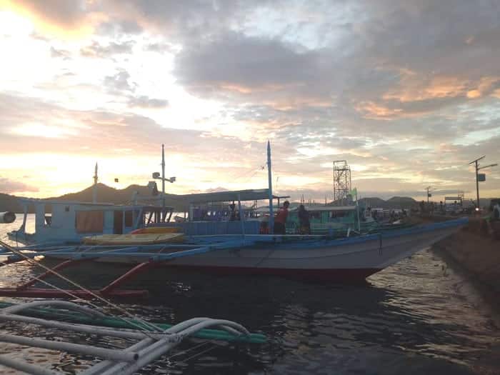 A colorful boat docked by the shore at sunset, with a cloudy sky painted in warm hues, invites thoughts of island hopping in the Philippines. Other boats and distant islands are visible in the background, creating a peaceful maritime scene.