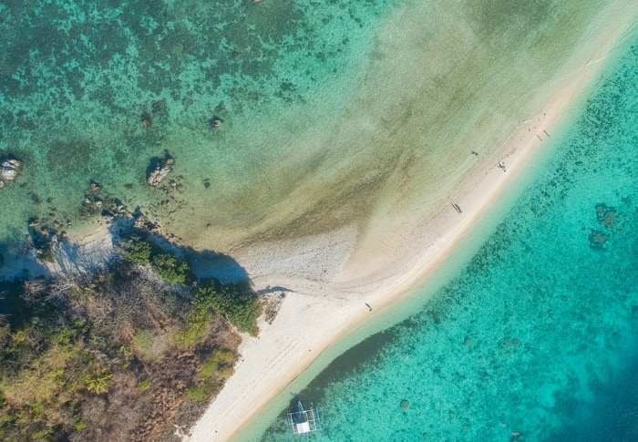 Aerial view of a narrow sandbar surrounded by clear turquoise water and patches of vegetation. A small boat from a nearby boat tour is docked near the sandbar, inviting explorers.
