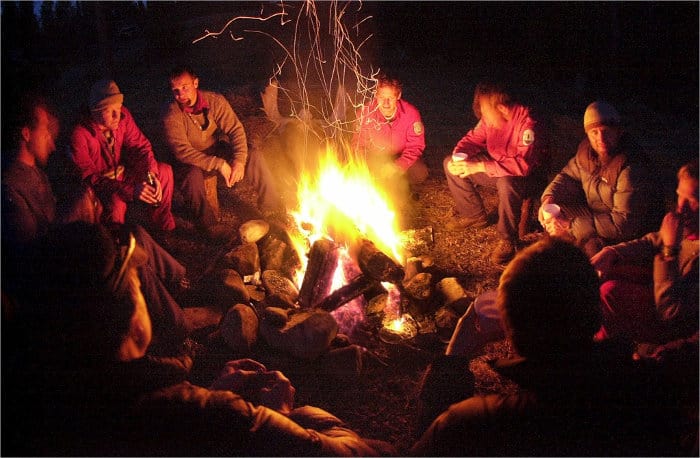 A group of people sitting around a campfire at night, warmly lit by the flames, surrounded by darkness, shared stories of their earlier boat tour.