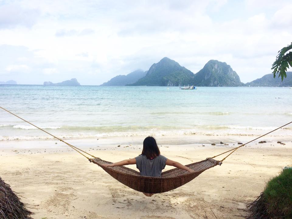 A person is relaxing on a hammock by a tropical beach in El Nido, with mountains and a boat tour gliding through the background.