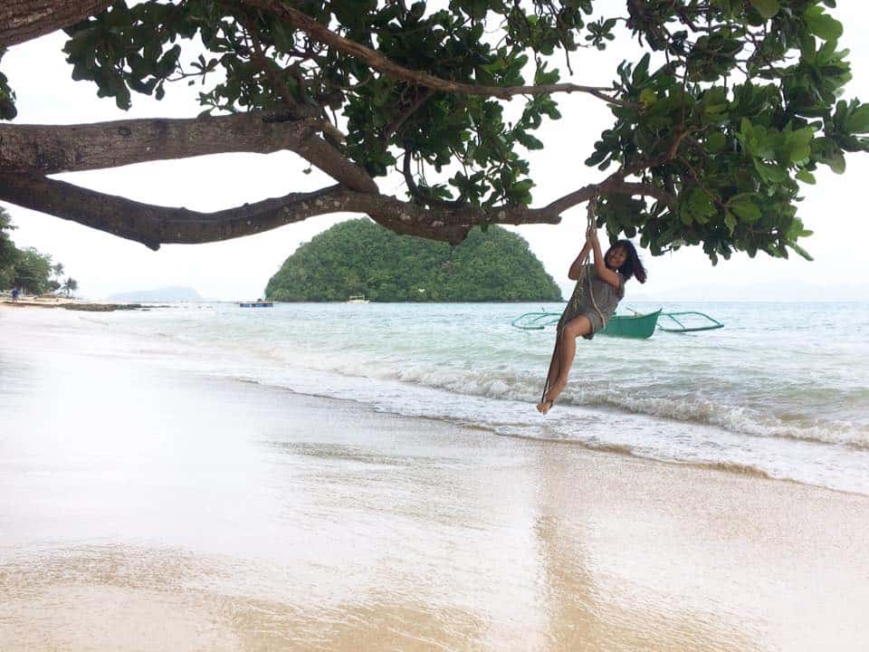 A person swings from a tree over a sandy beach, with the picturesque El Nido island gracing the background, as if inviting them to explore its beauty through a serene boat tour.