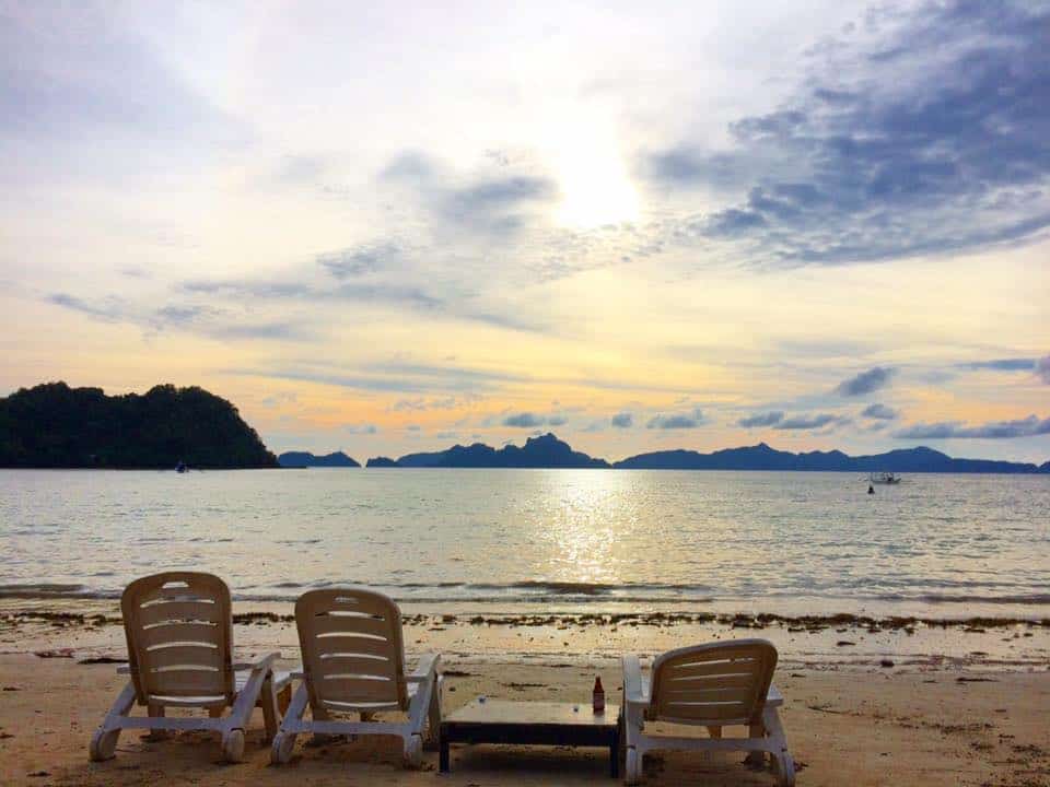 Three beach chairs facing a calm sea at sunset, with scattered clouds and distant islands reminiscent of an El Nido boat tour on the horizon.