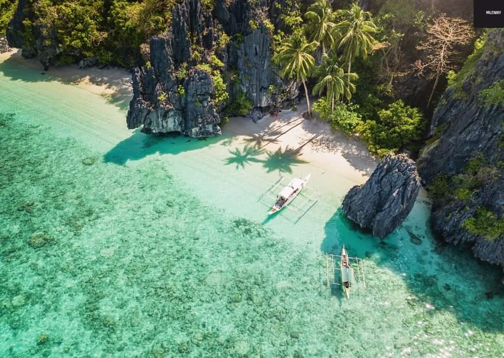 Aerial view of El Nido's tropical beach with clear turquoise water, rocky cliffs, lush palm trees, and boats ready for an unforgettable boat tour.
