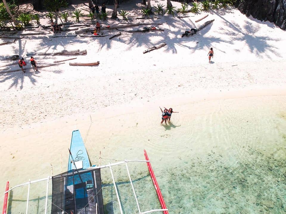 Aerial view of a tranquil El Nido beach with clear water, a boat from the local tour gliding peacefully, a few people onshore, and palm tree shadows casting on the sand.