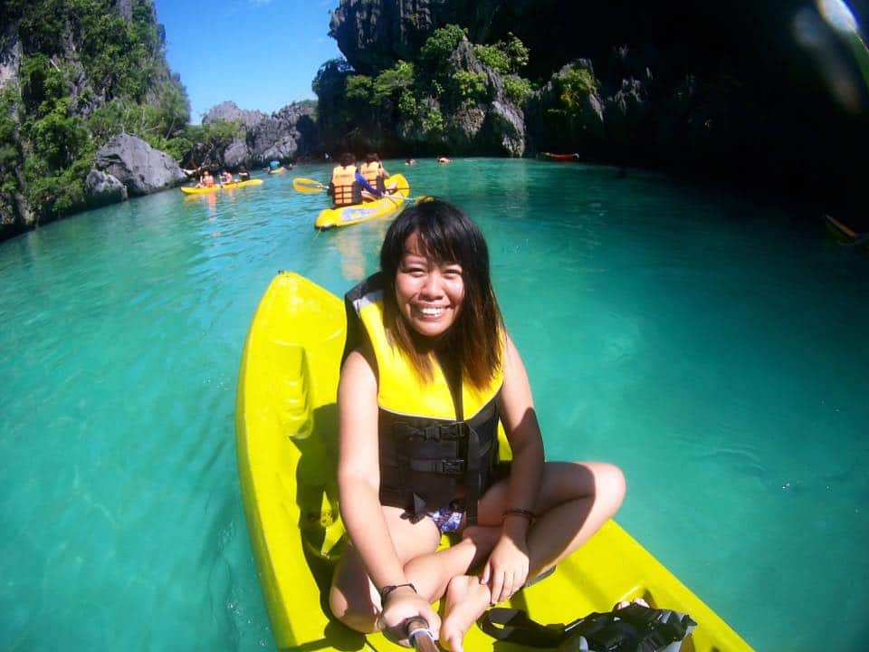 A person smiling on a yellow kayak in the clear turquoise waters of El Nido, surrounded by fellow kayakers and rugged rocky cliffs, offers an unforgettable boat tour experience.