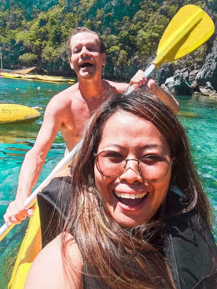 A smiling woman and man enjoy a kayaking adventure on a sunny day, navigating the clear blue waters of El Nido. They are surrounded by lush greenery and rocky cliffs, creating a picturesque backdrop for their unforgettable boat tour.