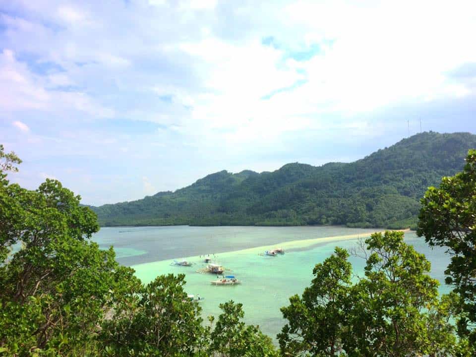 Scenic view of El Nido's bay with clear turquoise water, small boats awaiting a boat tour against the backdrop of lush green hills and a partly cloudy sky.