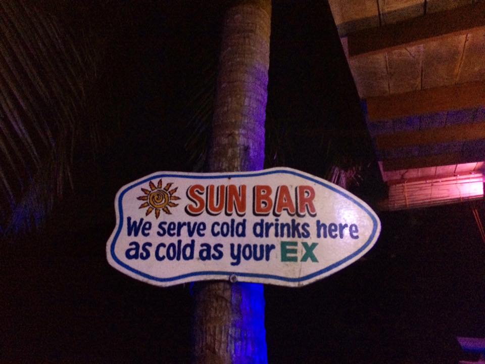 A sign reading "Sun Bar: We serve cold drinks here as cold as your ex" is attached to a palm tree at night, perfect for those returning from an El Nido boat tour.