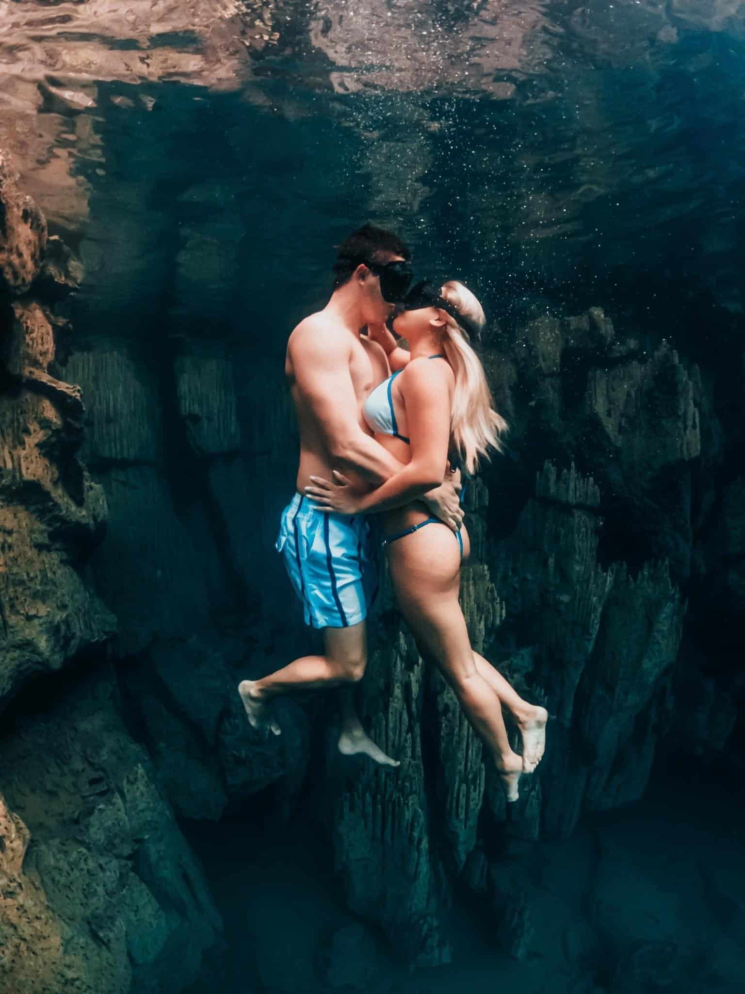 A man and a woman in swimwear kiss underwater, wearing snorkeling masks and fins amidst the clear waters of Palawan. Surrounded by rocky formations, the lighting creates a serene and romantic atmosphere, perfect for a unique marriage proposal.