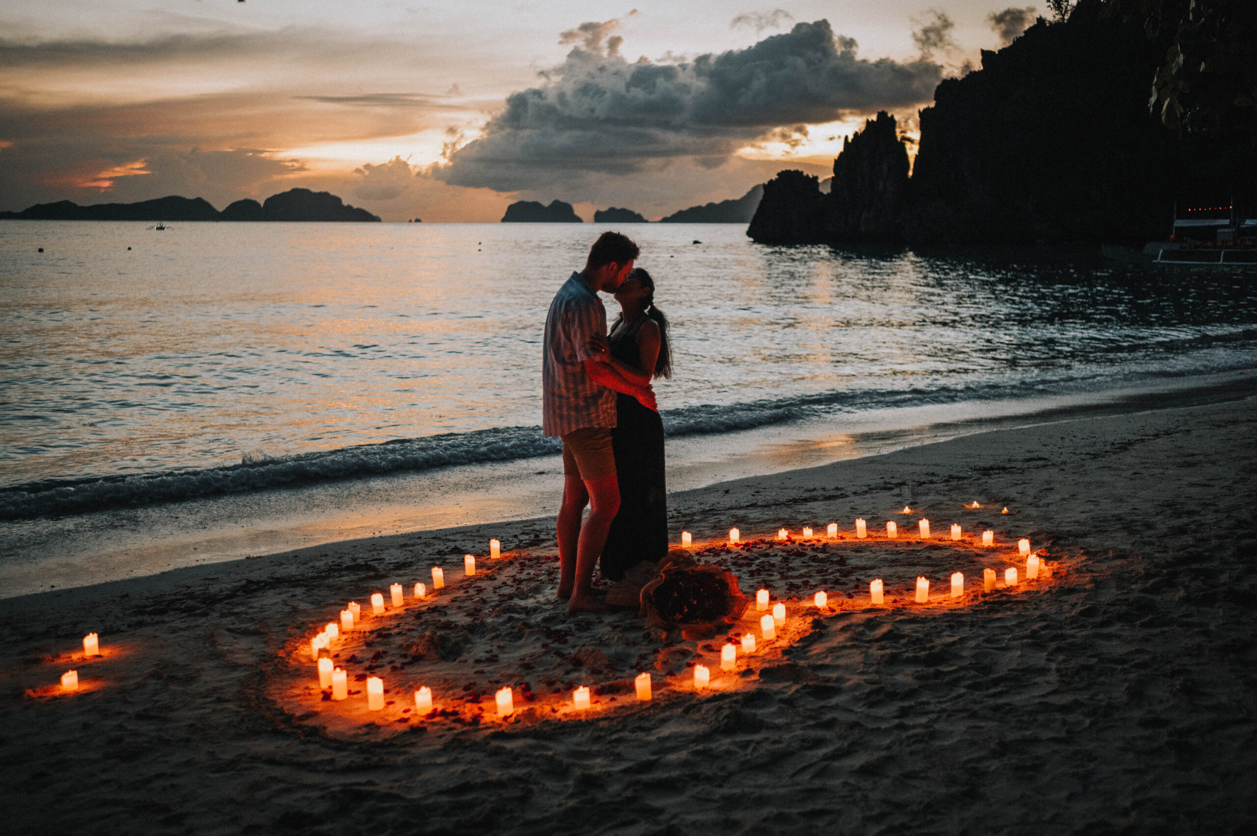 A couple embraces on a beach at sunset in Palawan, surrounded by candles in a heart shape—a dreamy proposal moment.