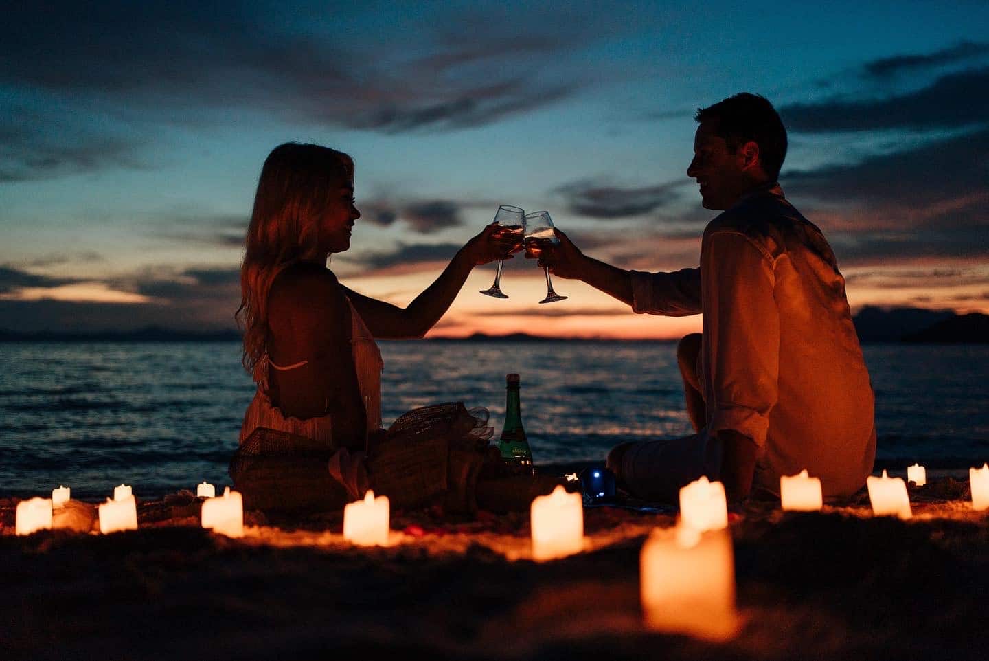 A couple sitting on a beach in Palawan at sunset, surrounded by lit candles, clinking wine glasses. The ocean and a colorful sky create a romantic atmosphere perfect for a marriage proposal.