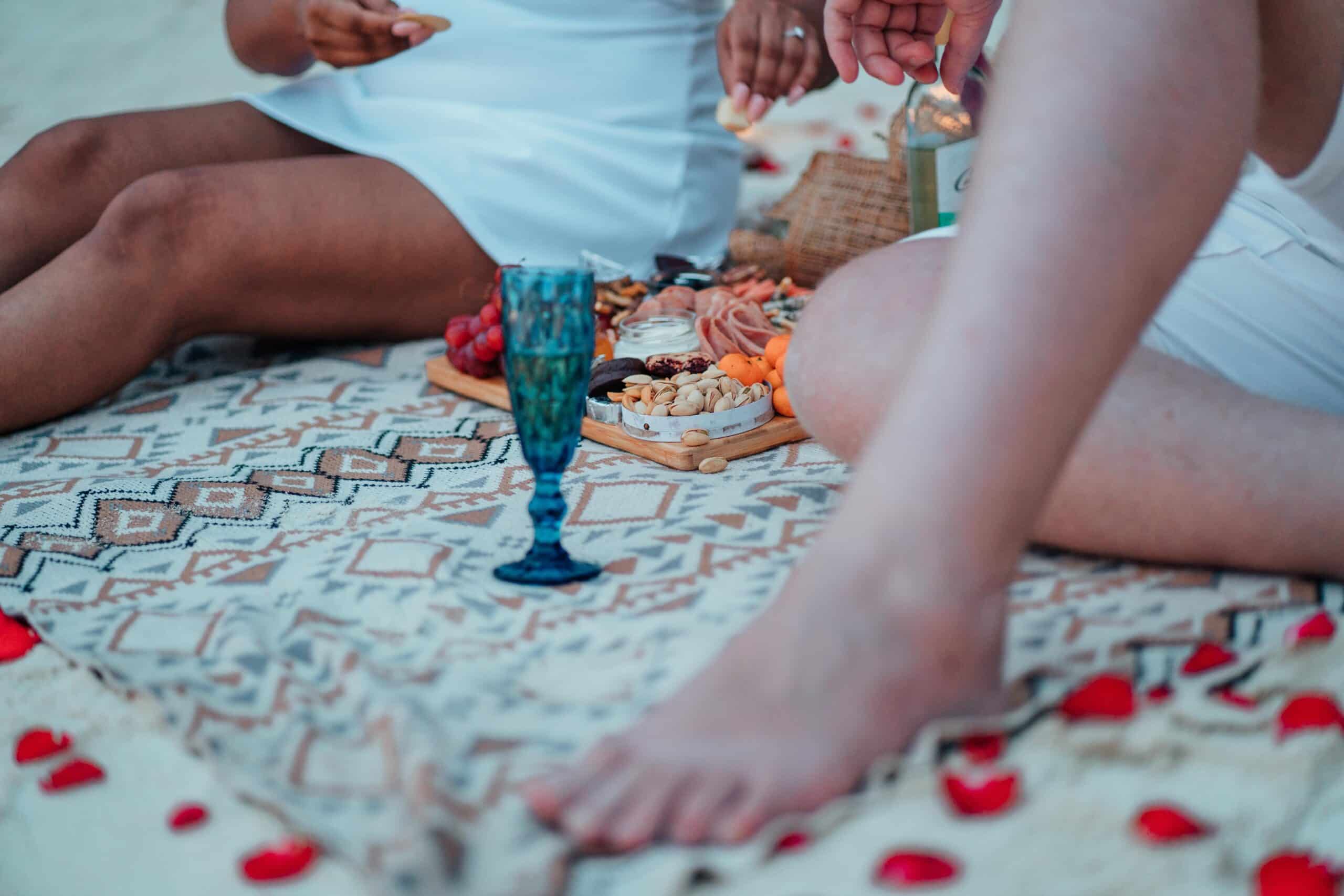 A romantic picnic on the beach features a patterned blanket with scattered rose petals. Two people, partially visible, savor a charcuterie board and drinks. A blue goblet is in the foreground, setting the scene for what could be a memorable marriage proposal after their Palawan boat tour.