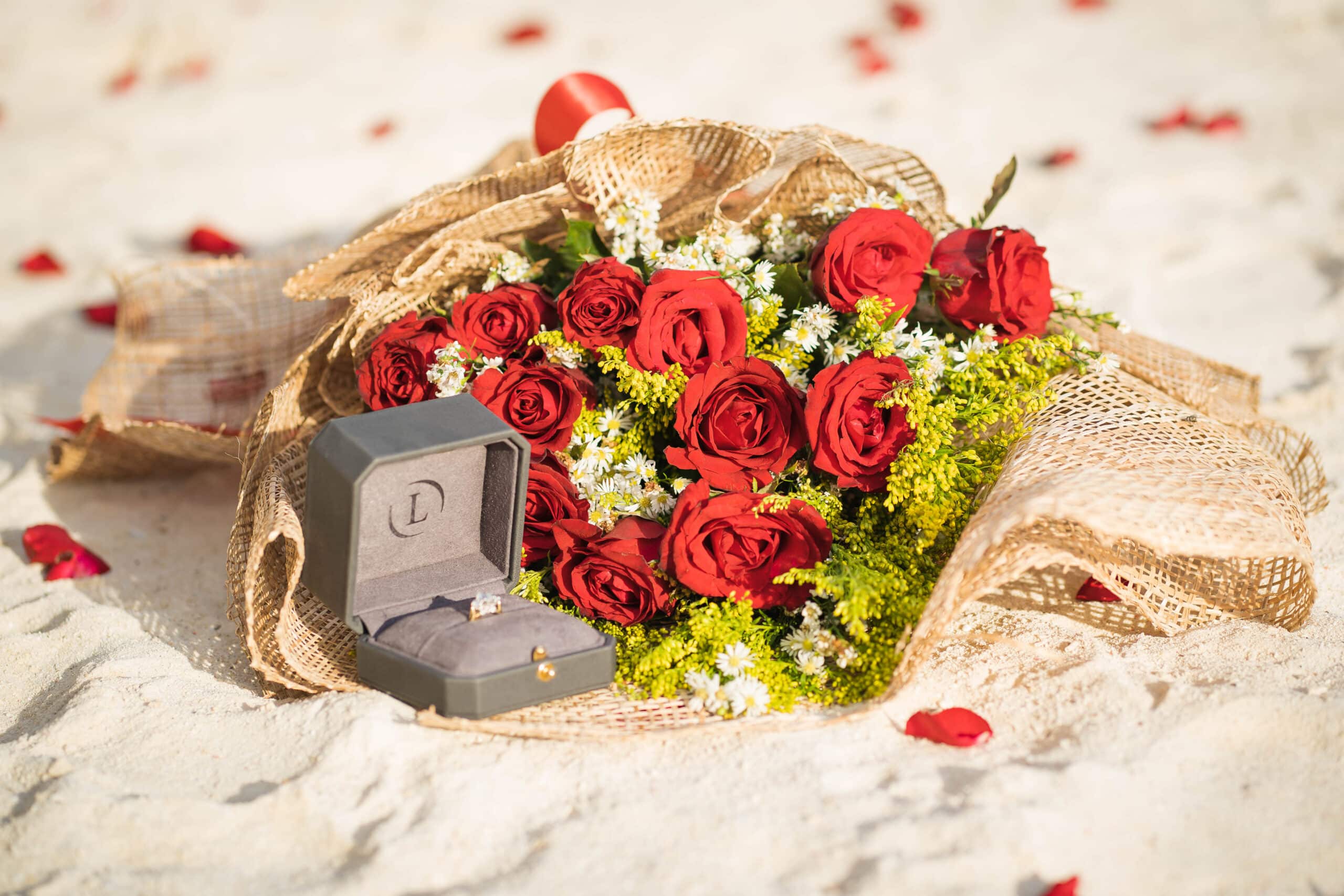 A bouquet of red roses and white flowers wrapped in burlap rests on sandy ground, the perfect setting for a marriage proposal. An open dark gray jewelry box with a ring inside is placed beside the bouquet, surrounded by scattered rose petals gently dancing in the breeze.