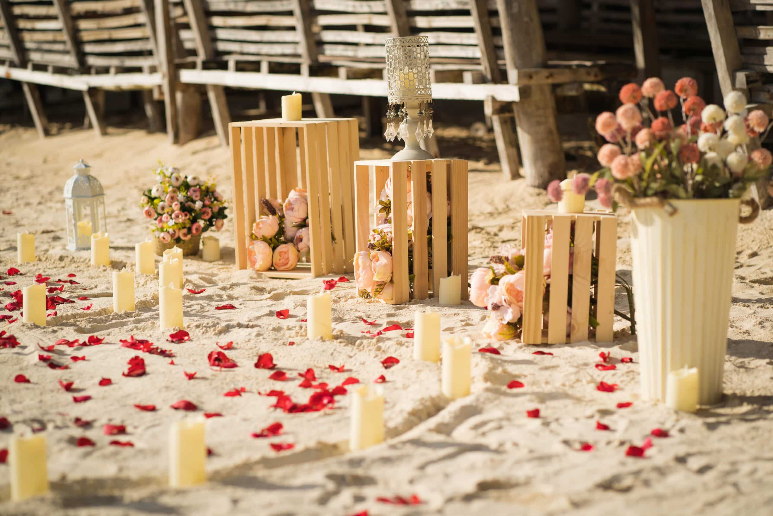 Beach setting on Seven Commando Beach with flower arrangements in wooden crates and vases, surrounded by numerous white candles. Red rose petals are scattered on the sand. A large lantern is also present, enhancing the romantic atmosphere of this idyllic El Nido locale.