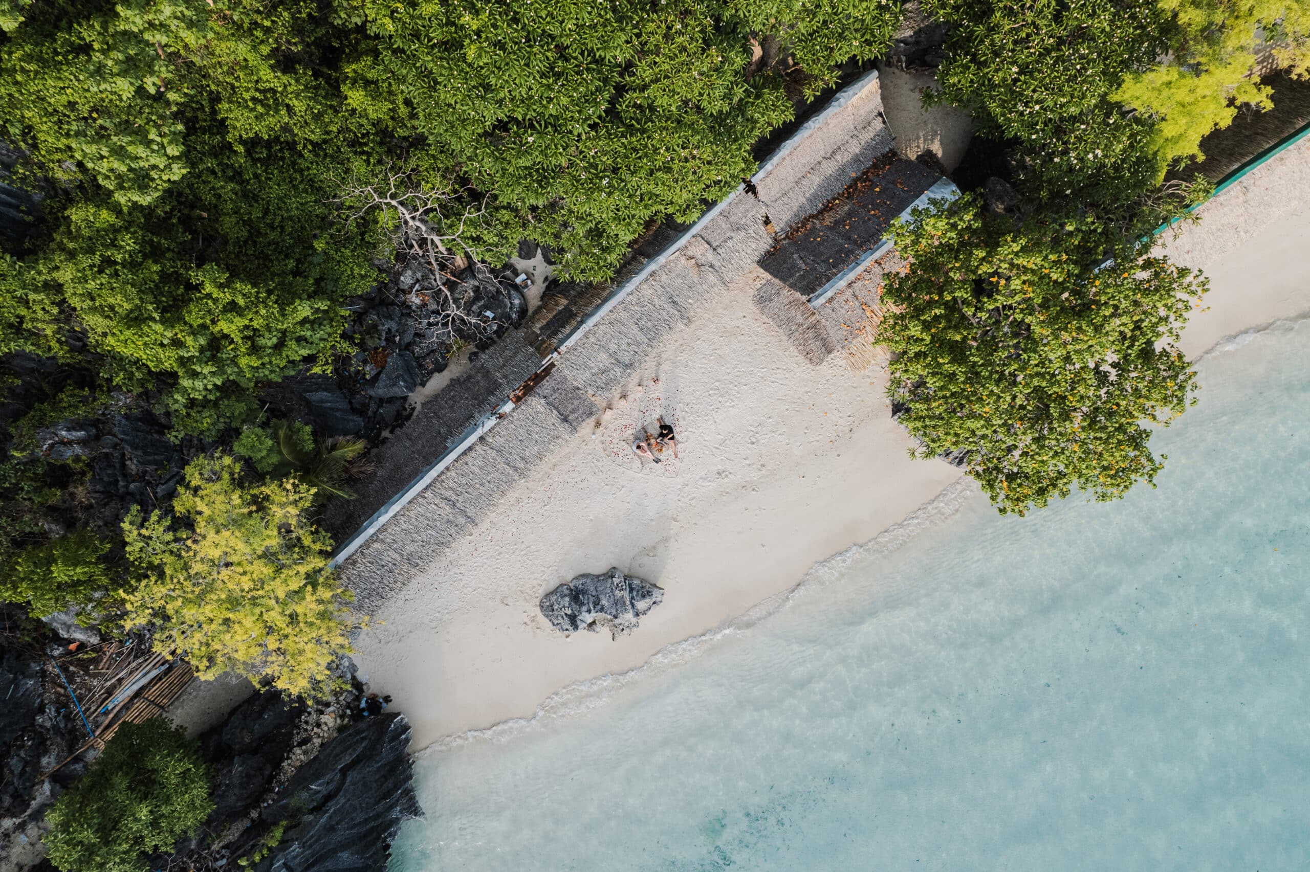 Aerial view of Seven Commando Beach in Palawan, where a small sandy shore is embraced by lush greenery. Two people sit on the sand, next to a large rock, as the clear blue waves of El Nido gently lap the shore.