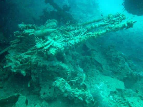 Underwater image of a shipwreck encrusted with marine growth, nestled in the vibrant reefs near Coron. The wreck is partially covered in aquatic plants and algae, resting amongst the ocean floor. The scene is illuminated with a blue-green tint from the surrounding water, perfect for scuba diving exploration.