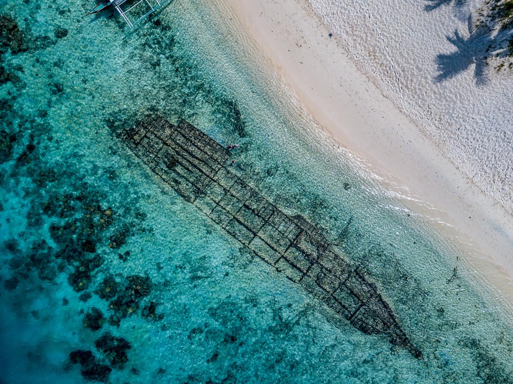 Aerial view of a shipwreck submerged in clear turquoise waters near a sandy beach. The ship's outline is visible beneath the shallow water, nestled among vibrant reefs and rocks. Palm tree shadows are cast on the sandy beach above, making it an inviting spot for scuba diving exploration.