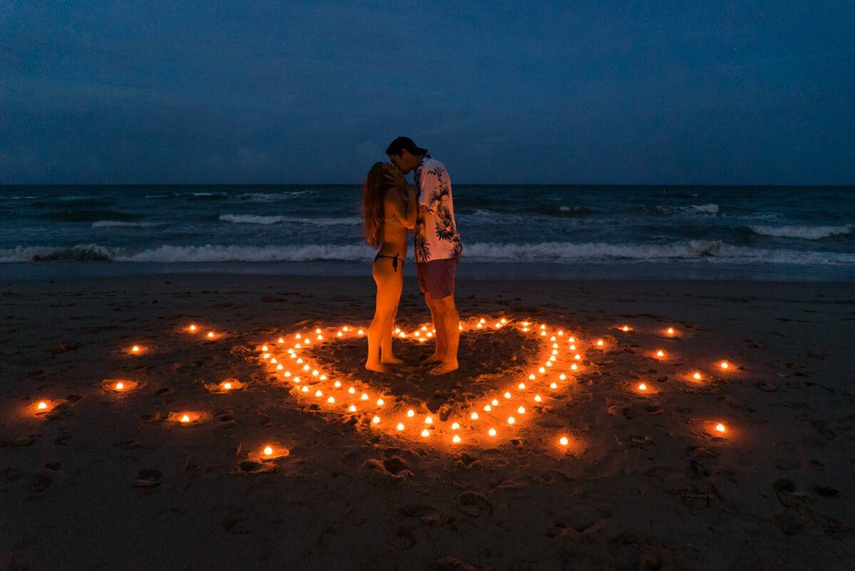A couple stands embraced on a beach in Palawan at night, surrounded by candlelight arranged in a heart shape on the sand, as the ocean waves gently roll in. It's the perfect moment for a marriage proposal under the starlit sky.