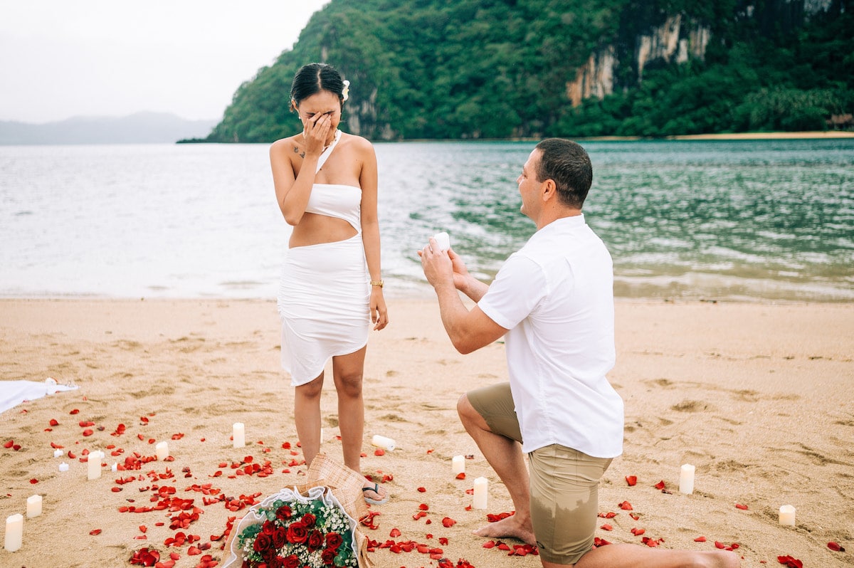 On a sandy beach in Palawan, adorned with red rose petals and candles, a person kneels holding a ring box, proposing to another who looks surprised and covers their face. Lush greenery from an island hopping tour and gentle waves create the perfect romantic backdrop.