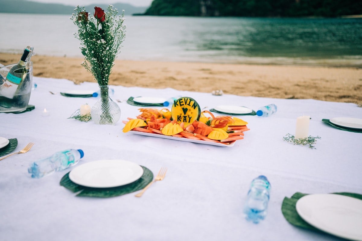 A beachside table setup on white cloth showcases plates and water bottles. A centerpiece displays tropical fruits and a carved watermelon with "Kevin & Kay" on it, hinting at a romantic marriage proposal. A vase with a red rose, greenery, and a candle completes the scene against the backdrop of Palawan’s ocean and hills.