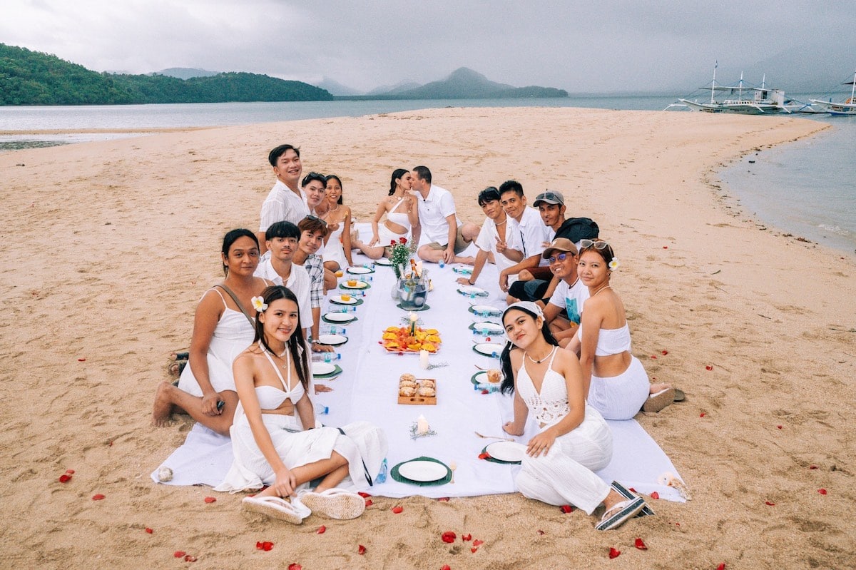 A large group of people dressed in white sit around a long table set for a meal on a sandy Palawan beach. The ocean and mountains are visible in the background, creating a perfect setting for what appears to be a marriage proposal celebration. A large group of people dressed in white sit around a long table set for a meal on a sandy Palawan beach. The ocean and mountains are visible in the background, creating a perfect setting for what appears to be a marriage proposal celebration.
