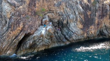 Along the rocky cliffside with wavy patterns above the blue sea, two people are climbing near small caves, a perfect spot on their island hopping adventure.