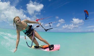 A person kiteboarding on clear turquoise water under a blue sky with scattered clouds, perhaps enjoying an exhilarating break during an island-hopping adventure.