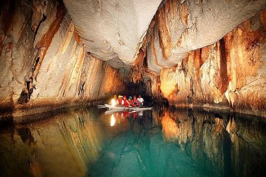 A small boat tour with people explores an underground river inside a large cave, showcasing textured, colorful rock walls and a calm water surface.