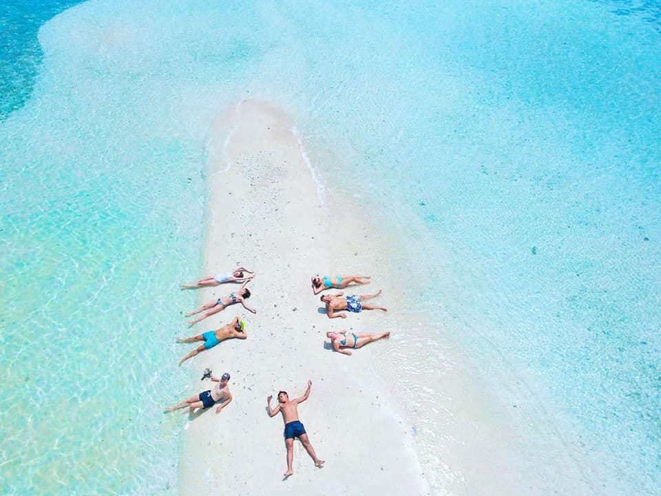 A group of people lying on a narrow sandy strip surrounded by clear turquoise water, enjoying the sun in this tropical paradise at Big Lagoon, Palawan. The vivid blue shades of the ocean contrast with the bright sand, creating a serene and picturesque scene perfect for island hopping adventures.