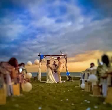 A couple exchanges vows under a decorated arch with a sunset sky backdrop, surrounded by seated guests on a grassy lawn. The ceremony marked the perfect conclusion to their island hopping adventure, with stories of each boat tour echoing in their hearts.