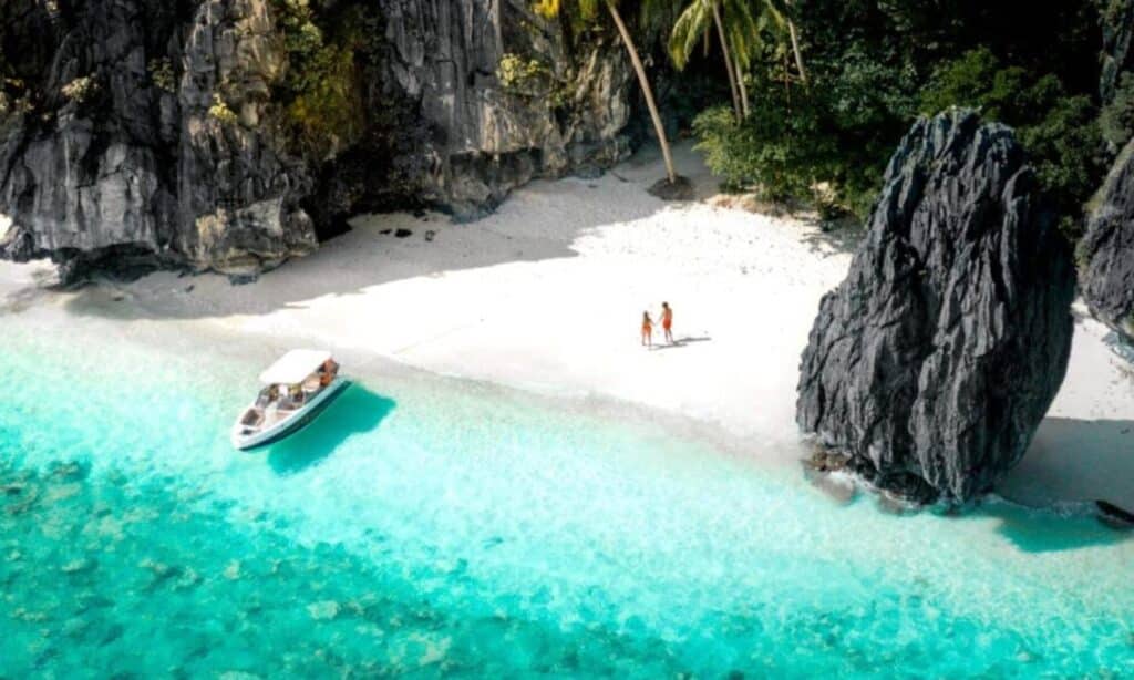 A boat moored near the tropical beach of Coron basks in the turquoise water beside a rocky cliff and swaying palm trees, setting the scene for an unforgettable expedition. Three people stand on the white sand, soaking up the idyllic surroundings.