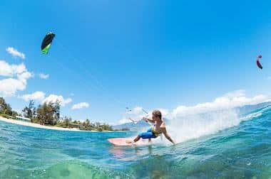 A person is kiteboarding on clear blue water near a tropical beach, possibly taking a break from island hopping under a bright blue sky with a few clouds.
