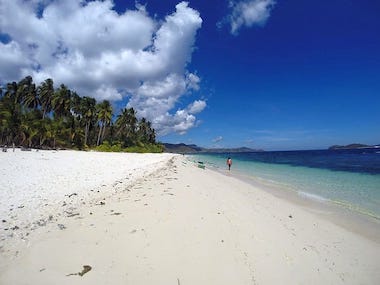 A tranquil beach with white sand, clear turquoise water, and a person walking along the shore. Palm trees line the beach under a bright blue sky, perfect for island hopping adventures.
