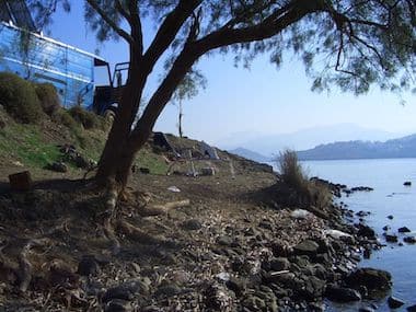 Tree and rocky shoreline beside a lake, perfect for island hopping, with a distant view of mountains and a blue truck partially visible on the left.