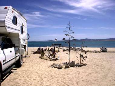 A camper van is parked on a sandy beach with a view of the ocean under a clear blue sky. Rocky formations and a small pier hint at upcoming boat tours for adventurous island hopping in the background.