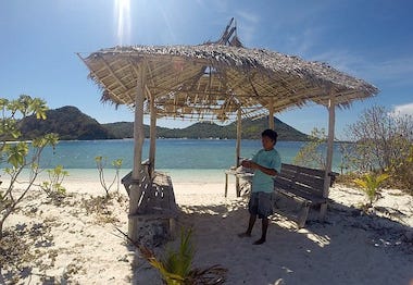 A person stands under a rustic thatched hut on a sandy beach, with clear blue water and green hills in the background, dreaming of island hopping adventures or perhaps taking a boat tour to explore hidden gems.