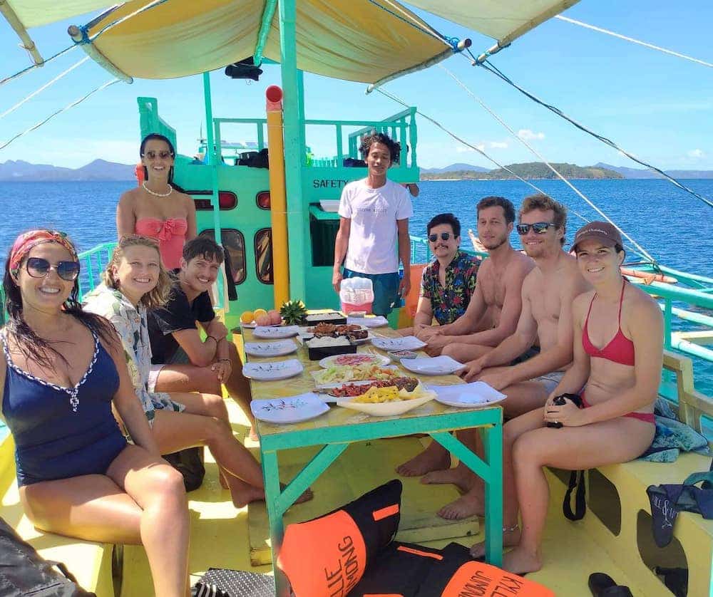 A group of people in swimwear sits and stands around a table brimming with food on a boat, under a canopy. The serene waters and distant islands of El Nido form the backdrop to this exciting expedition.
