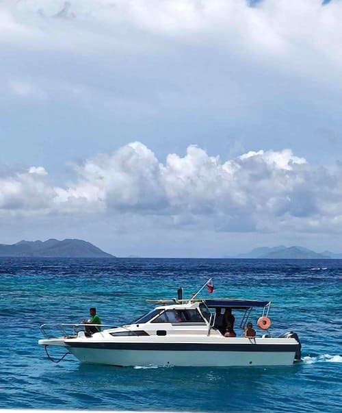 A small white motorboat with people on board is island hopping through clear blue waters. In the background, distant islands and a cloudy sky create a picturesque scene. The boat, equipped with a lifebuoy and a flag at the stern, gracefully glides along its adventure.