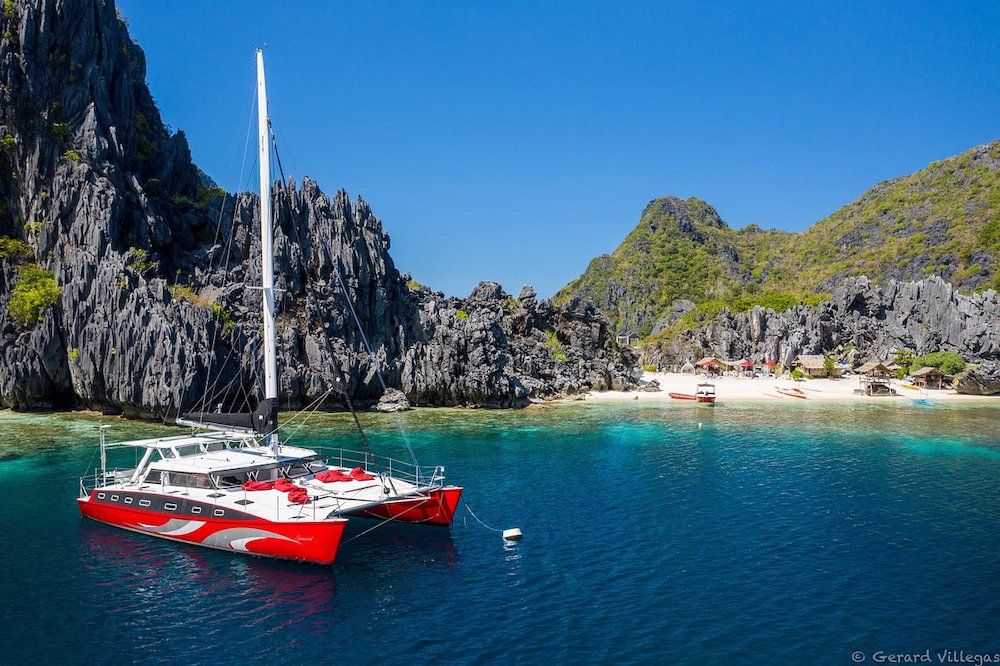 A vibrant red catamaran floats on clear blue waters near a small sandy beach with jagged cliffs covered in green vegetation, perfect for island hopping in the Philippines. Several small boats of various sizes are docked near the shore under a bright blue sky.