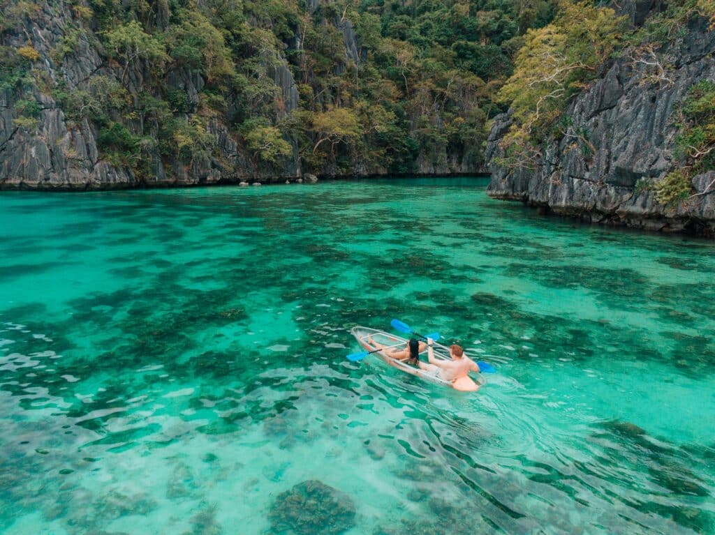 Two people embark on an exhilarating kayaking excursion in Coron, gliding through clear turquoise waters surrounded by lush, rocky cliffs and vibrant greenery.