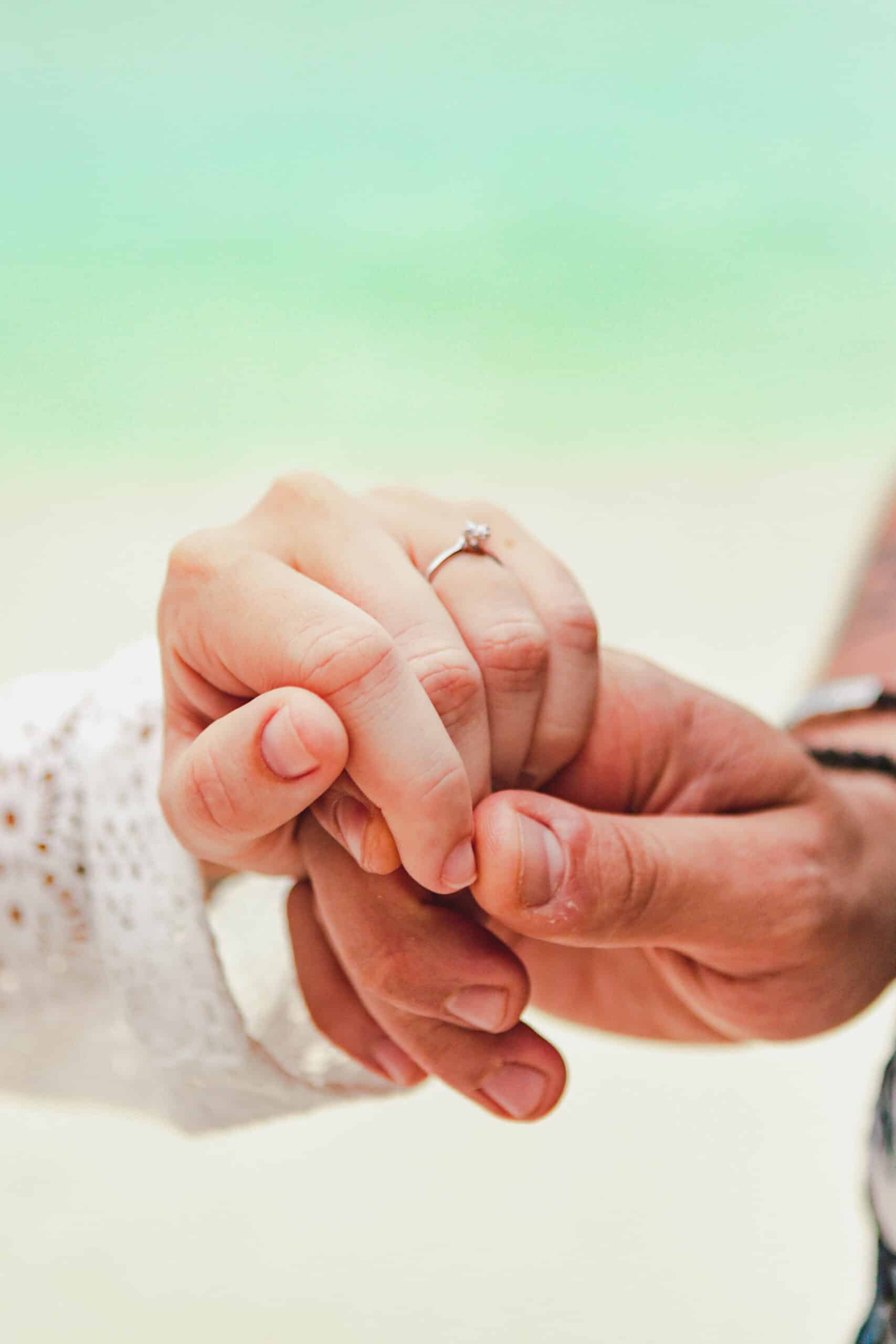 Close-up of two hands holding each other, one adorned with a diamond engagement ring. The romantic background is blurred, showcasing soft colors reminiscent of a beach vacation or ocean setting.