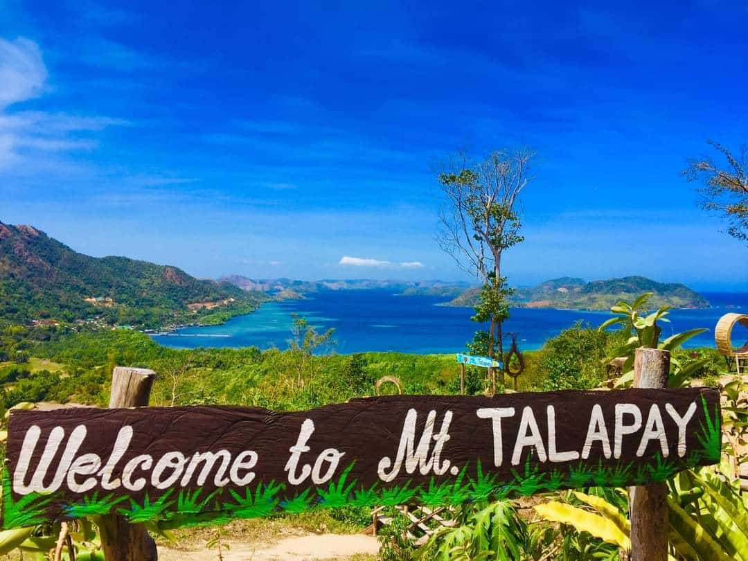 Scenic view from Mt. Talapay with a wooden sign in the foreground reading "Welcome to Mt. Talapay." In the background, lush green hills and a vast blue ocean reminiscent of Coron are visible under a clear sky, inviting thoughts of an idyllic boat tour adventure.