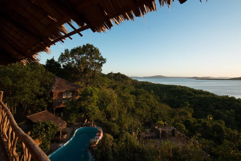 A serene view from a thatched roof overlooking a lush, green jungle with a turquoise pool nestled among the foliage. The scene, reminiscent of El Nido, extends to a calm, expansive body of water under a clear blue sky at dusk.