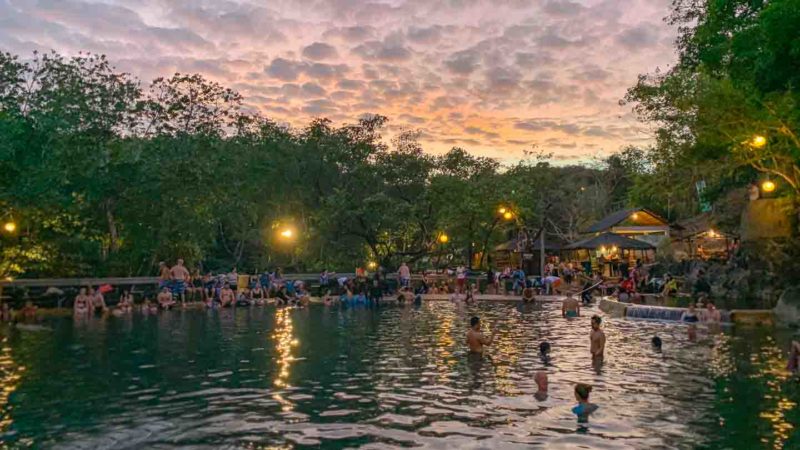 People enjoying a natural hot spring pool at sunset, reminiscent of El Nido's serene escapes. The sky is filled with clouds, and trees surround the area. The warm lighting from nearby lamps creates a cozy atmosphere as visitors relax in the water, much like after an adventurous boat tour.