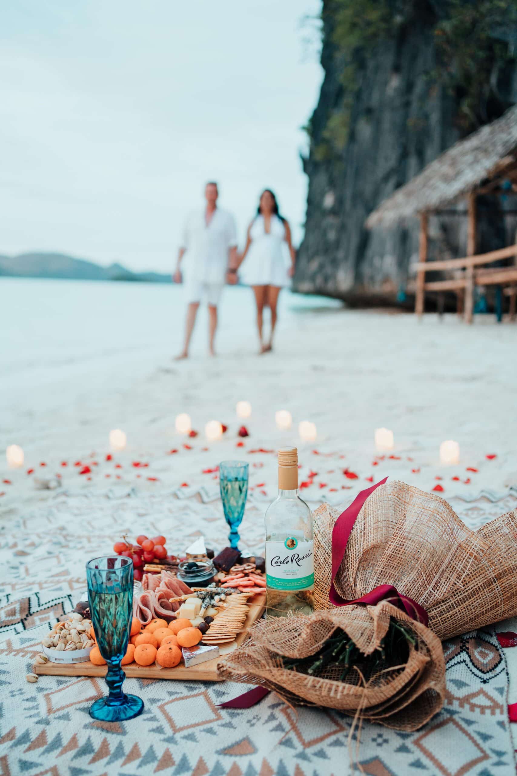 A romantic beach picnic in the Philippines features a blanket with assorted fruits, cheese, and a bottle of wine. Two people stroll along the shoreline as candles and rose petals adorn the sand. Nearby, thatched huts and cliffs complete this idyllic destination.