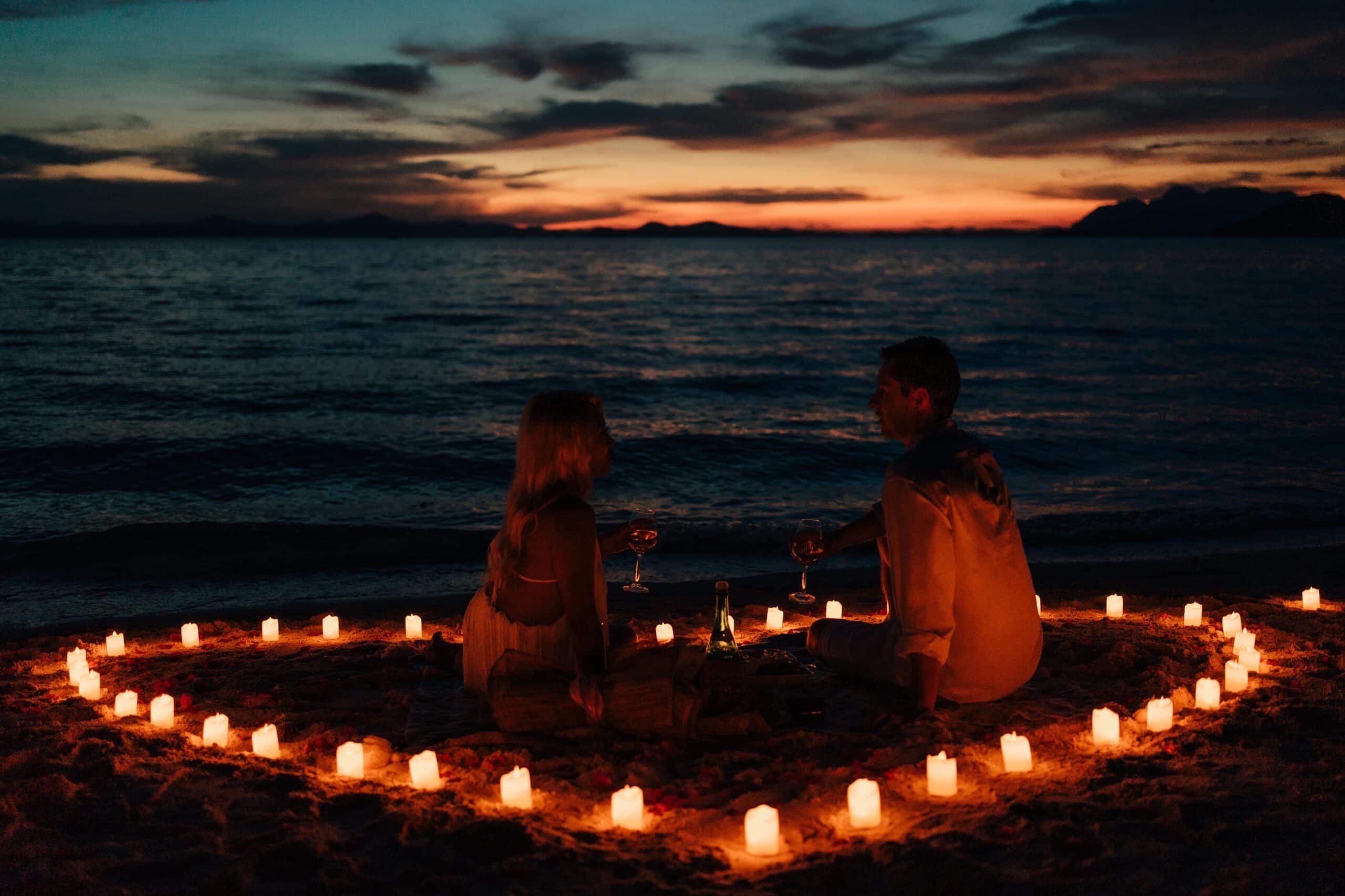 A couple sits on El Nido's Seven Commando Beach at sunset, embraced by a heart-shaped arrangement of candles. Facing the ocean with glasses in hand, they watch as the Palawan sky transitions from orange to deep blue.