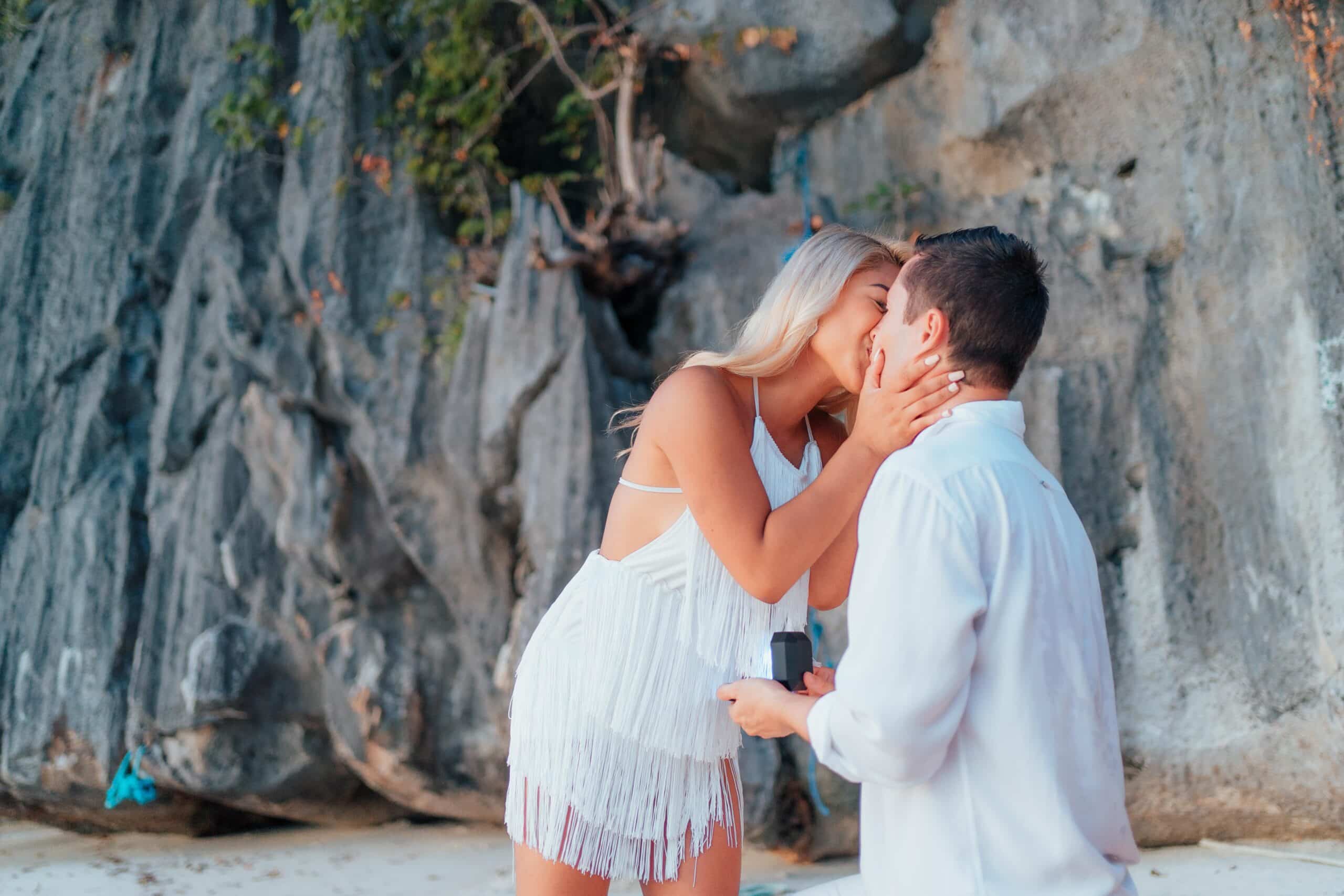 A couple shares a romantic kiss on a rocky beach in the Philippines. The woman in a white dress leans down to kiss the man who is kneeling and holding a small box, suggesting a proposal against the backdrop of rocky cliffs and lush foliage—a perfect moment from their vacation.
