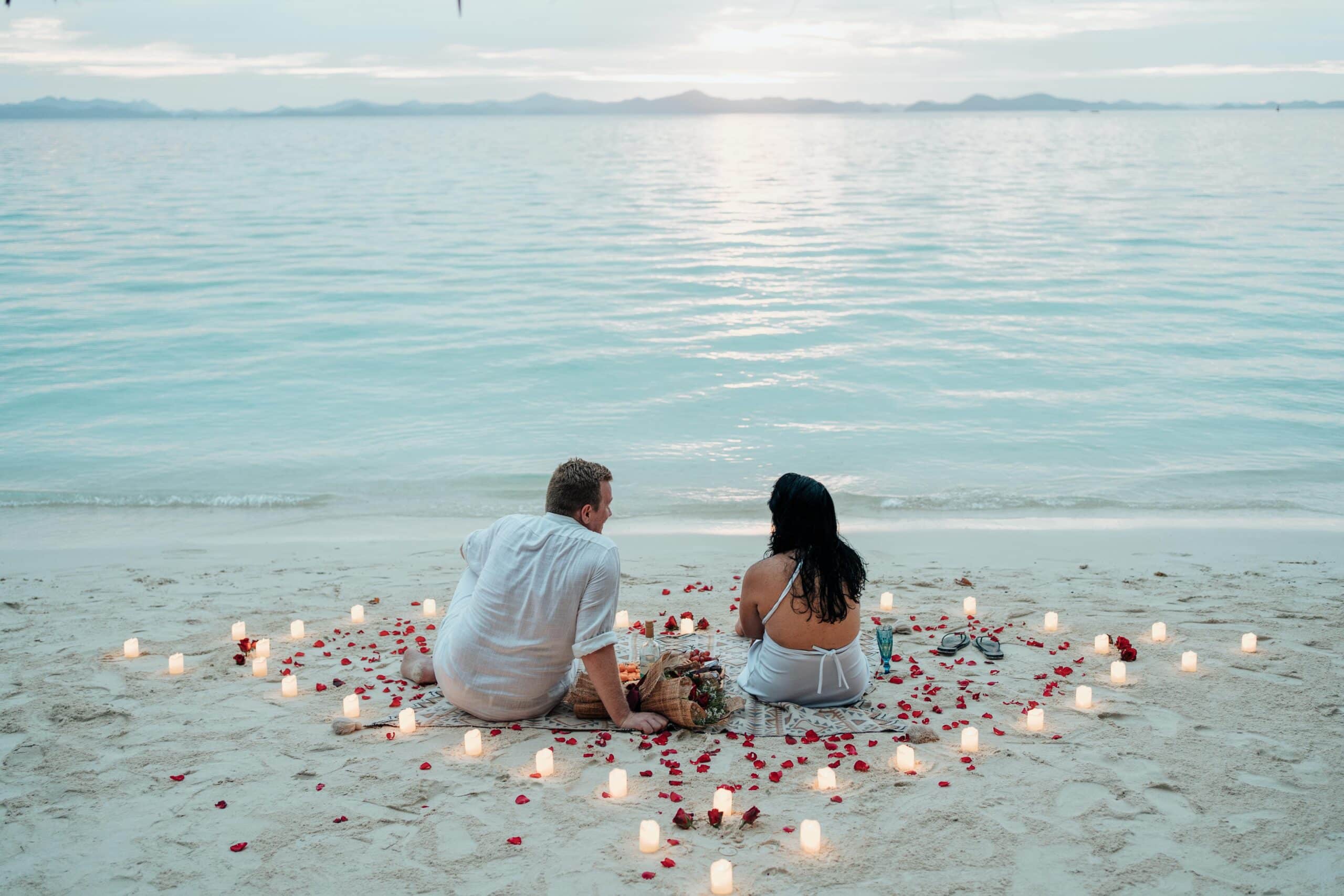 The couple sits on a beach surrounded by candles and rose petals, gazing at the ocean's sunset, reminiscent of their unforgettable island hopping adventure in Coron.